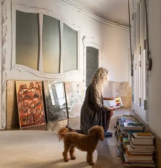 Viladomiu examines some of the books she has stored in her magnificent apartment. Artworks stand propped-up against the wall behind her, while her dog stands attentively beside her.