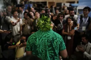 Back of Sikulu wearing green shirt and headpiece as he faces audience