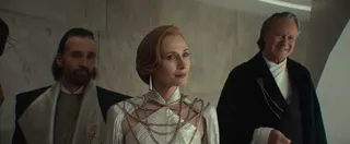 A shot of Andor season 2. It features Perrin Fertha, a smug-looking human male with a goatee and pushed back brown hair. He wears fancy garb. Next to him is Mon Mothma, who wears a white, crystal-like dress with fancy earrings. She sports short, pushed back ginger hair and looks serious. To her right is Luthen Rael, wearing a black and white fancy robe. He looks pleased as punch.