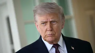 Close-up of President Trump  with gray hair in a dark suit, white shirt, and red tie, wearing a small American flag pin; he has a serious, furrowed expression.