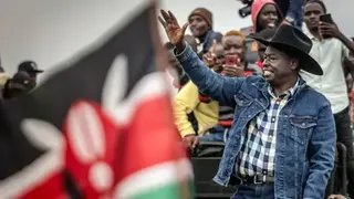 AFP via Getty Images Rigathi Gachagua wearing a denim jacket, checked shirt and black cowboy hat waves and smiles to supporters. He is on the right of the picture. In the foreground, on the left, is a Kenyan flag out of focus.