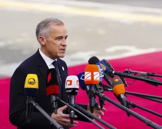 Carney, standing on a red carpet speaks into a large array of microphones