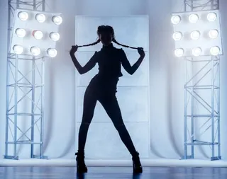 Beautiful slim young woman with two braids dressed in a black clinging clothes. The young woman dancer is performing the dance in the vogue style. She is standing holding the braids in hand. Studio shooting on a white background with light blue backlighting on a two stands