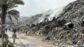 A mountain of debris steams in the heat on a street in Gaza. It's as high as the buildings and people walk by. Since the start of the war, most of Gaza's infrastructure has been destroyed.