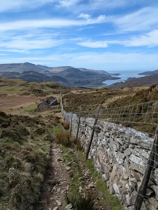 A view back down a stony track by a drystone wall from the side of a mountain in Wales, with other mountains and the sea in the distance.