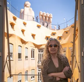 Ana Viladomiu, the last tenant in Gaudí’s apartment building Casa Míla (better known as La Pedrera), stands on her balcony. Behind her, the distinctive, yellow exterior of Casa Milà’s inner block is bathed in sunshine.