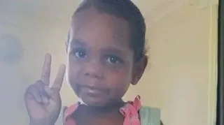 Northern Territory Police An Aboriginal girl wearing a pink dress and blue backpack poses for a picture, holding up the peace sign