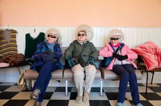 three elderly women sit on vintage chairs on a black and white floor with what appear to be plastic bowls for hairdrying on their heads. They wear anoraks and black sunglasses and are laughing. There are hand-knitted tank tops hanging on the wall behind them.