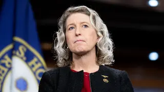 Portrait of a woman with blue eyes and wavy blonde hair, wearing a red top and black textured jacket with a gold pin; a blue and yellow flag blurred in the background.