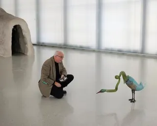 A man with thinning white hair kneels down on the floor of a gallery to look closely at a sculpture of a long-necked bird-like creature.