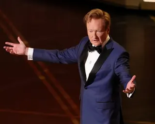 man in tuxedo speaks on stage while holding arms out