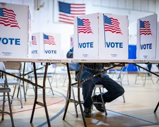 A person sits at a voting booth with privacy dividers displaying American flags and the word 'VOTE'.