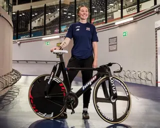 Katie Archibald stands beside a black track bike at the National Cycling Centre in Manchester.