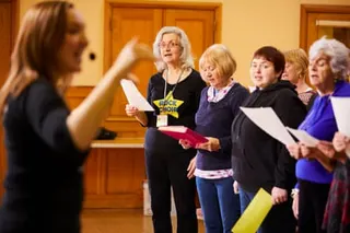 Women of varying ages, but mostly older and with grey hair, sing in a choir as a young woman conducts them.