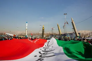 A series of rockets and missiles pointed into the sky among a group of people and a large Iranian flag.