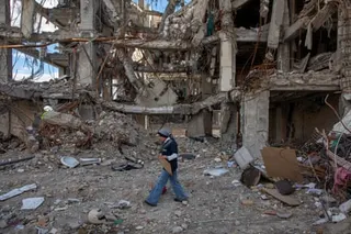 A person in a vest walks through rubble and debris in front of a heavily damaged multi-story building.