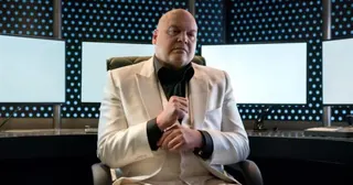 heavyset bald men in white suit over black shirt, sitting authoritatively in an ultramodern office