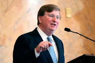 A man in a dark suit and blue striped tie gestures while speaking at a podium.