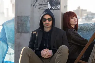 man in dark glasses and hoodie seated at the base of a column with a woman in a red wig off to the side
