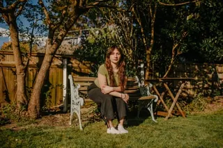 Adele Zeynep Walton sits on a bench in a sunny garden with her elbows on her knees, leaning forwards. She is in her mid-20s with long brown hair and wears a green T-shirt.