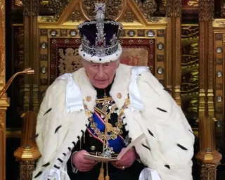 King Charles III in ceremonial robes and crown reads from papers while seated on an ornate throne