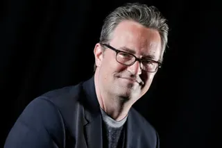 Matthew Perry in glasses and a dark blazer smiles slightly against a black background.