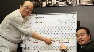 一張照片顯示兩名男子指著一幅裝框的元素週期表中的第113號元素，該元素旁標有日本國旗