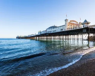 Brighton Palace pier with ornate buildings extends over calm sea water, with a pebble beach in the foreground.