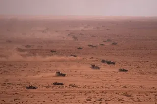 Military vehicles drive across a sandy desert terrain kicking up dust trails.