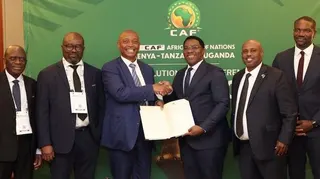 A group of four sports ministers and football bosses from Tanzania and Uganda, all wearing dark suits, white shirts and dark ties, pose for a photo alongside Confederation of African Football president Patrice Motsepe (third from left) and secretary general Samson Adamu (far right), who are similarly dressed. The group stands in front of a green board which features a Caf logo and Motsepe is shaking hands with one of the administrators as the pair hold open a large document for the camera