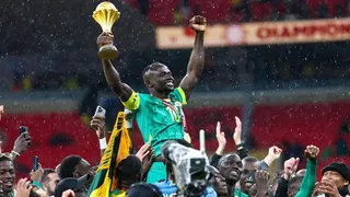 Senegal player Sadio Mane, dressed in green kit and wearing a golden medal on a red ribbon around his neck, is held aloft by fellow players as he raises both arms into the air, holding the Africa Cup of Nations trophy in his right hand.