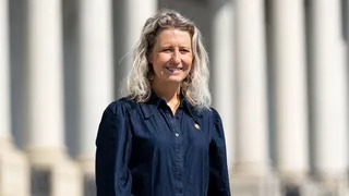 Smiling woman with wavy gray-blond hair stands outdoors in front of a columned building, wearing a dark denim shirt and a brown belt.
