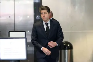 Sam Altman in a dark suit and tie walks through a modern building interior, glancing to the side