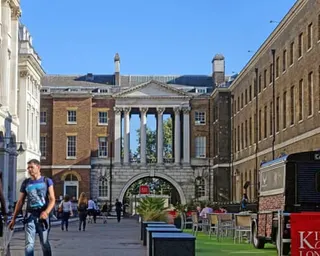 A classical stone archway with columns leads through a courtyard, with pedestrians and outdoor seating