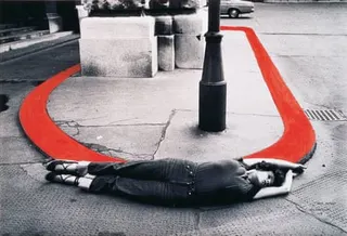 A black and white image of a raised oval section of pavement surrounded by road; in the foreground, a woman lies on her side with arms and legs extended, her body curved against the shape of the raised section; the kerb has been coloured red in post-production.