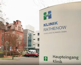 A white sign for Klinik Rathenow clinic with an arrow points to the main entrance, with buildings in the background.