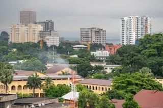 A Kinshasa cityscape with modern high-rise buildings, construction cranes, and lush green vegetation under an overcast sky