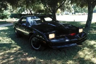 A black sports car with pop-up headlights, parked on grass under trees.