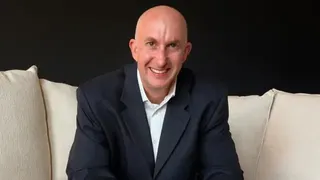 比斯利對著鏡頭微笑。他穿著深色西裝，坐在一張白色沙發上