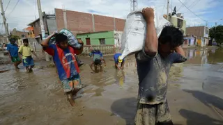 men walking through a flooded street covered in muddy flood water carrying food supplies on their shoulders.