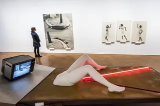 Works by Valie Export. In the foreground, Geburtenbett (Birth Bed): an outsized female abdomen with crooked legs on a mattress, red neon strip lights streaming from her vagina, and a TV transmitting a Catholic mass where the head would be. On the wall in the background hang a couple of Export's other works.