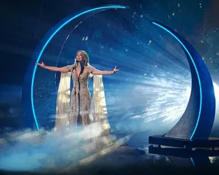 Delta Goodrem in a gold gown stands with arms outstretched on a misty stage beneath a large illuminated blue arch