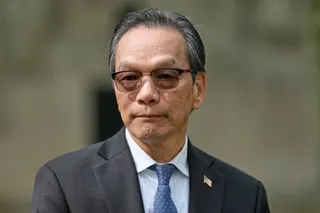 Lu Jianwang in a dark suit, glasses, and blue tie looks directly at the camera.