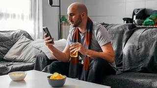 Young man looking at his phone while sitting on a sofa watching sports