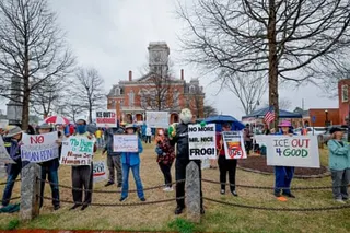 Protesters hold signs including 'ICE OUT' and 'NO MORE MR. NICE FROG!' in front of a historic courthouse building