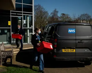 Two masked NHS workers carry red boxes to a van outside a modern building