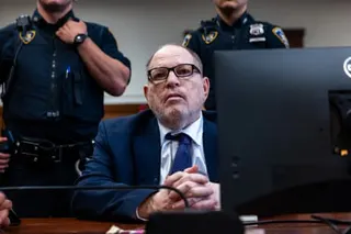 Harvey Weinstein in a suit and glasses sits at a courtroom desk with police officers standing behind him