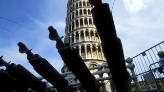 一排鑽探機械整齊排列在前景，正在進行工程作業；背景中可見比薩斜塔，隱約矗立於遠方，形成工程現場與歷史建築之間的對比