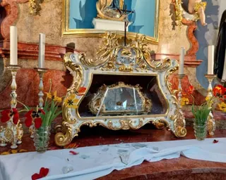 An ornate gold and white reliquary case sits open and empty, its glass smashed, on a red marble altar, flanked by candles and flowers