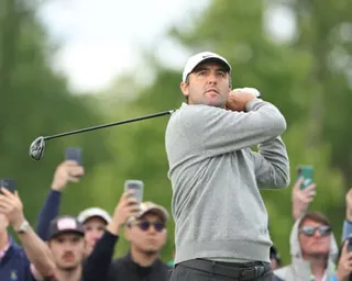 Scottie Scheffler in a gray sweater and white cap follows through on his swing while spectators watch